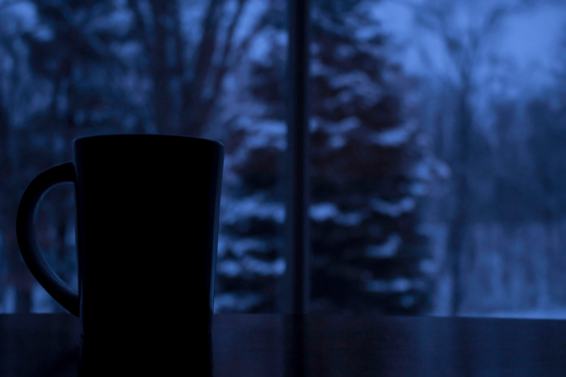 An image of a cup of hot chocolate on a table in front of a window. The outside is blue with dusk and snow.